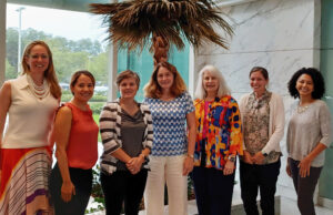 Seven women pose in front of a window.