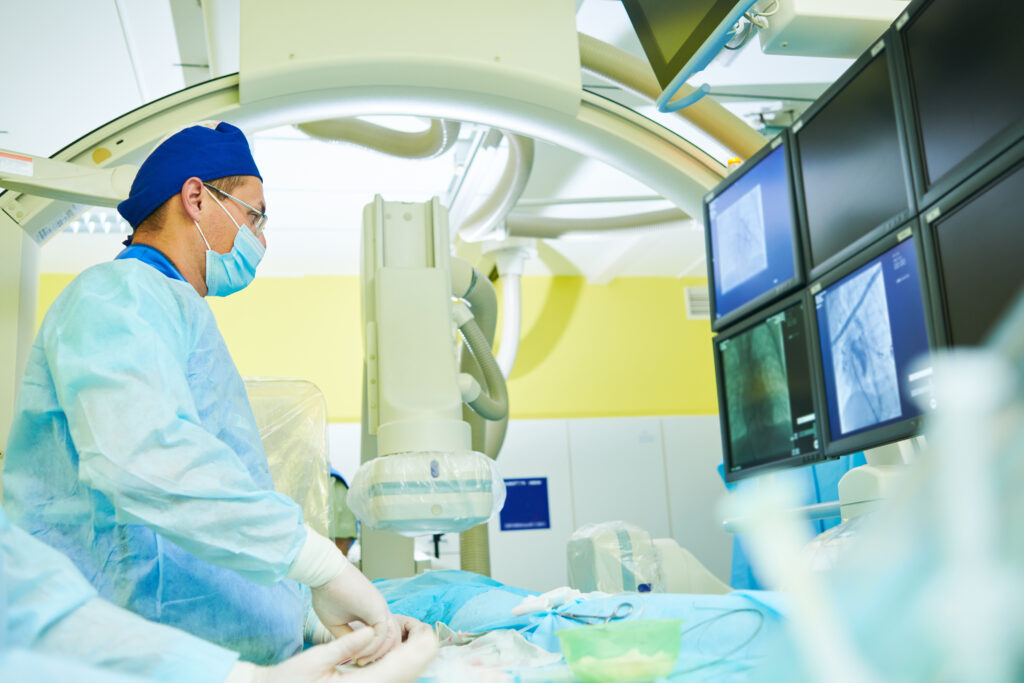 An interventional radiologist in face mask and scrubs stands over a patient and looks at images of lungs on a screen.