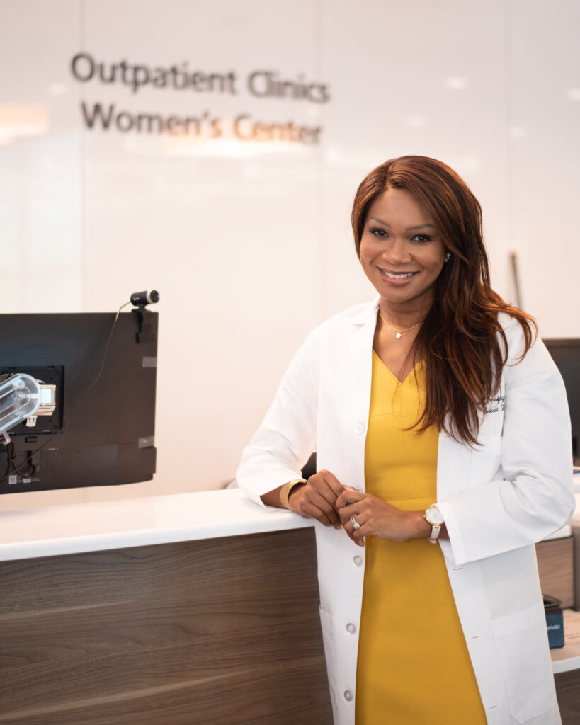 Dr. Omofoye, in a yellow dress and white lab coat, stands at a counter in the Outpatient Clinics Women's Center.