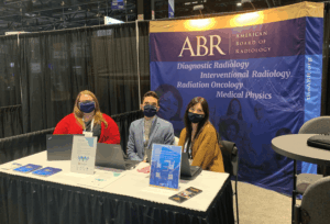Three ABR staff members sit at a table in front of a banner reading diagnostic radiology, interventional radiology, medical physics, and radiation oncology.