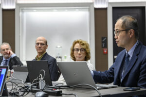 Three board members sitting at a table in front of laptop computers look at a fourth member who is speaking.