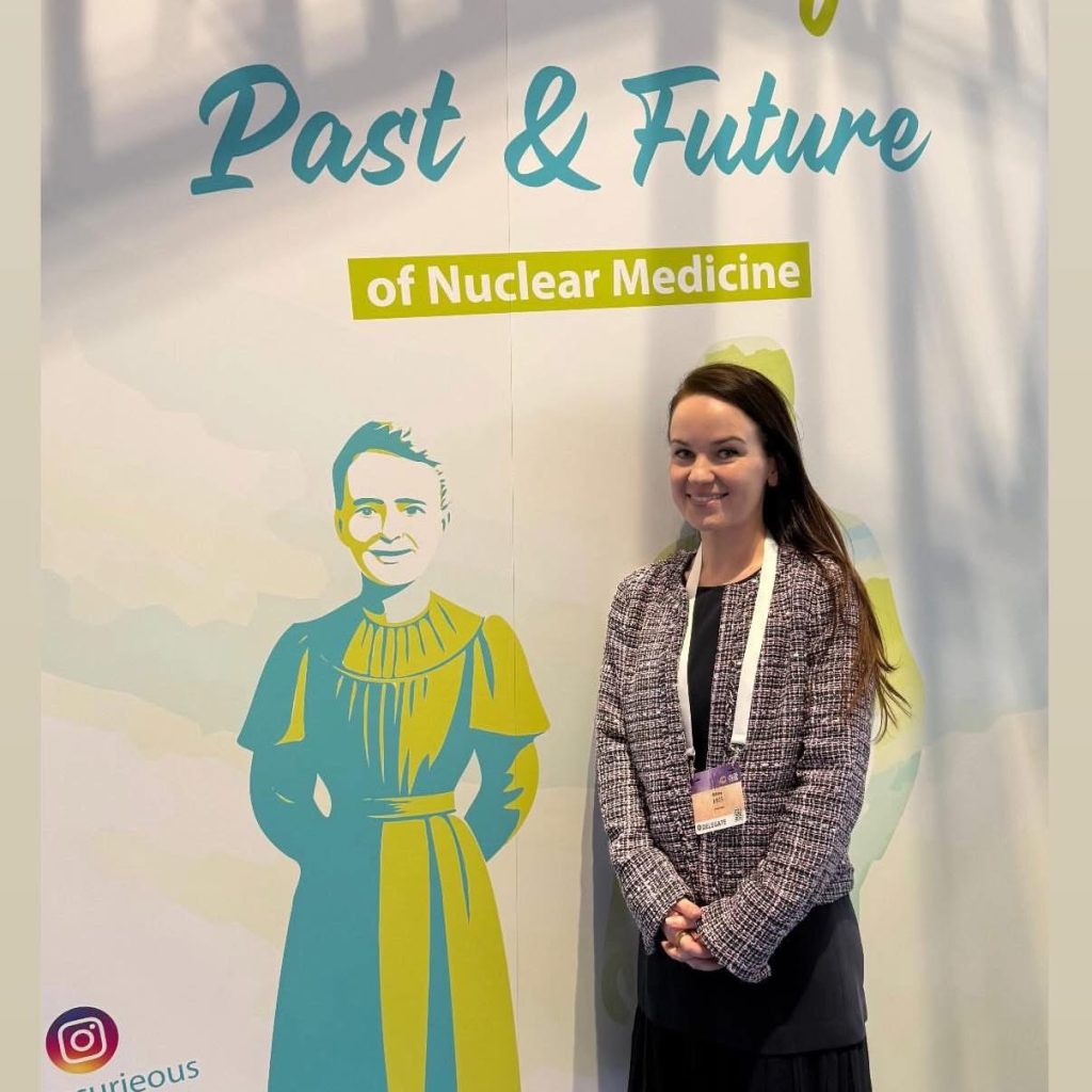 Dr. Miles stands in front of a poster labeled past and future of nuclear medicine with an image of Marie Curie on it.